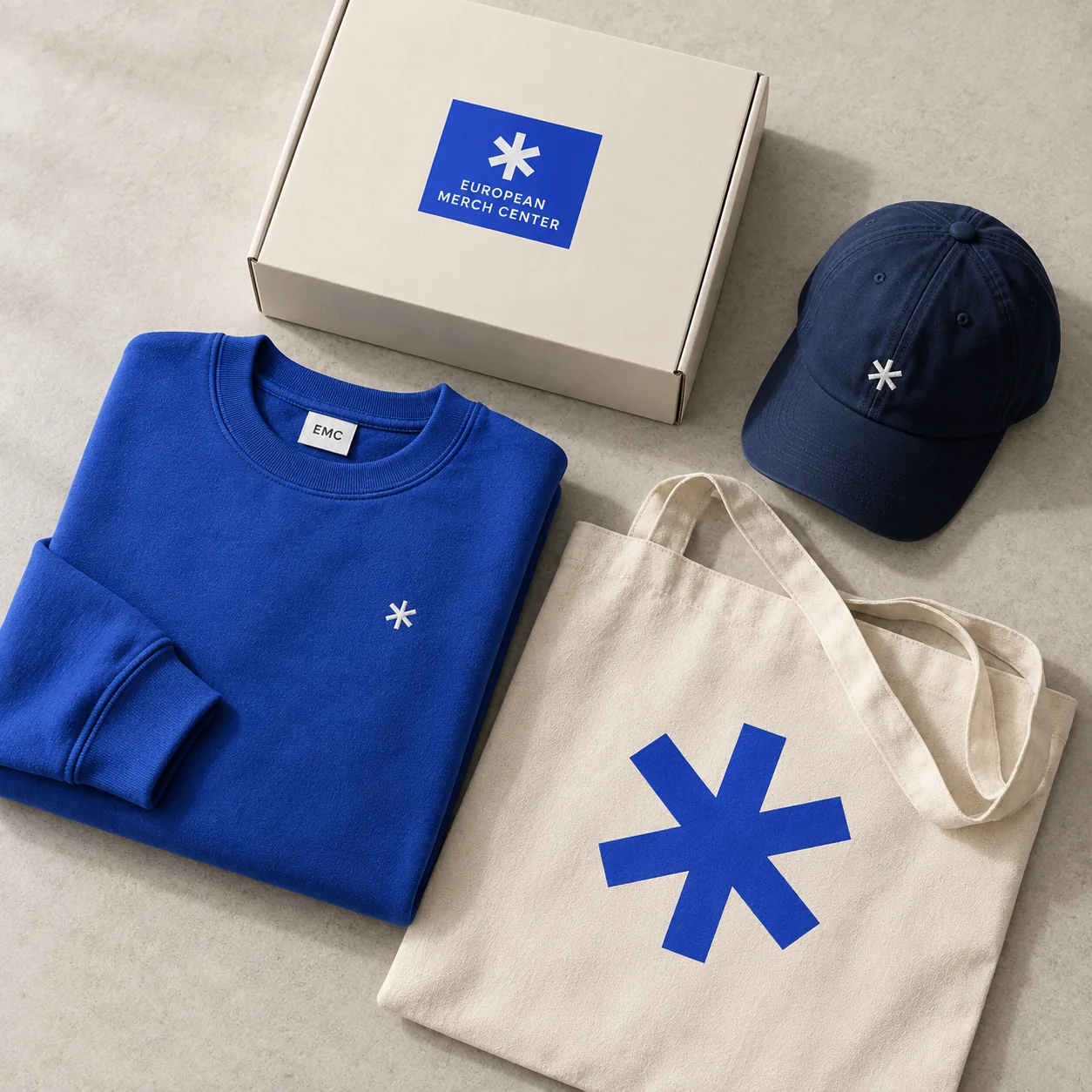 EMC-branded cobalt sweatshirt, ivory canvas tote, navy cap, and kraft gift box arranged on a warm gray surface